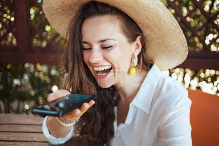 happy stylish middle aged housewife in white shirt with hat sitting at the table speaking on a smartphone in the patio.