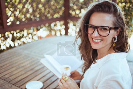 Foto de Retrato de mujer moderna sonriente en camisa blanca con taza de café y anteojos sentados en la mesa de lectura libro en la terraza del hotel casa de huéspedes. - Imagen libre de derechos