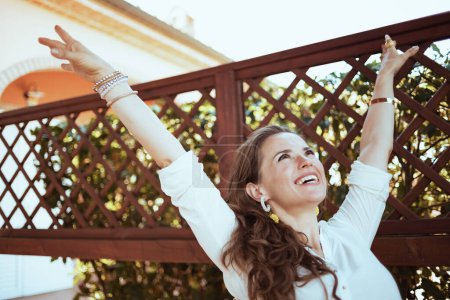 Foto de Feliz mujer moderna de 40 años de edad en camisa blanca en la terraza del hotel casa de huéspedes con los brazos abiertos regocijo. - Imagen libre de derechos