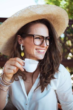 Foto de Sonriente ama de casa de mediana edad en camisa blanca con taza de café, sombrero y gafas en la terraza. - Imagen libre de derechos