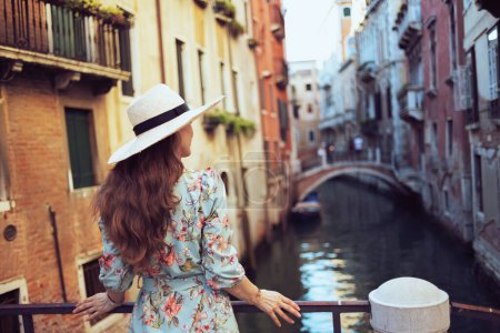 Photo for Seen from behind trendy woman in floral dress with hat sightseeing in Venice, Italy. - Royalty Free Image