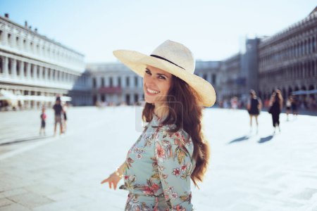 Foto de Mujer moderna feliz en vestido floral con sombrero explorar atracciones en la plaza de San Marco en Venecia, Italia. - Imagen libre de derechos