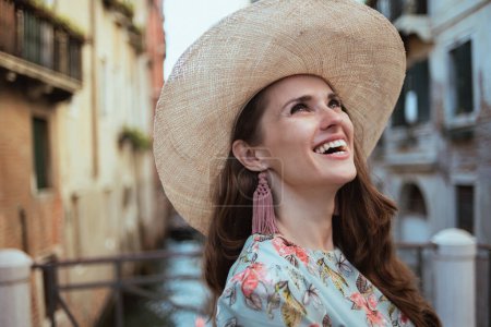 lächelnde stilvolle Frau im floralen Kleid mit Hut genießt die Promenade in Venedig, Italien.
