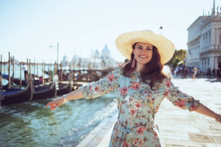 Photo for Smiling trendy woman in floral dress with hat having excursion on embankment in Venice, Italy. - Royalty Free Image