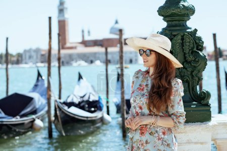 Foto de Mujer viajera moderna de mediana edad en vestido floral con gafas de sol y sombrero que tiene excursión en terraplén en Venecia, Italia. - Imagen libre de derechos