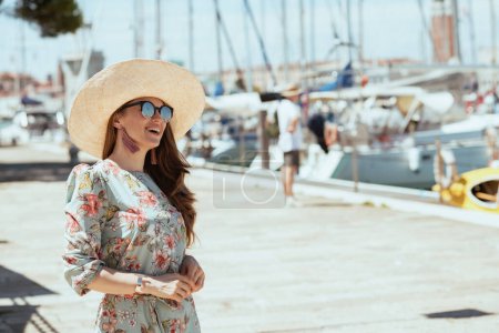 Foto de Mujer turista elegante sonriente en vestido floral con gafas de sol y sombrero en el muelle. - Imagen libre de derechos