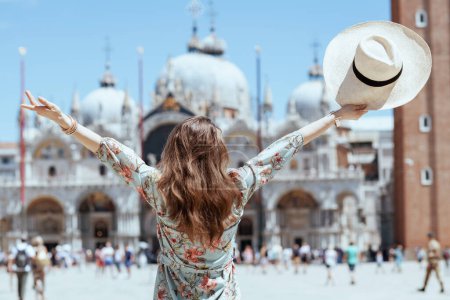 Foto de Visto desde atrás mujer viajera en vestido floral con sombrero en la plaza San Marco en Venecia, Italia con los brazos levantados regocijándose. - Imagen libre de derechos