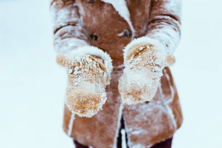 Photo for Closeup on middle aged woman in brown hat and scarf in sheepskin coat with snowy mittens outside in the city in winter. - Royalty Free Image