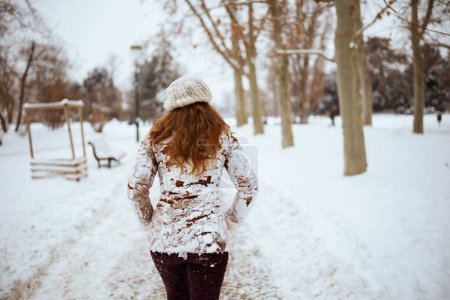 Photo for Seen from behind middle aged woman in brown hat and scarf in snowy clothes and sheepskin coat outside in the city in winter. - Royalty Free Image