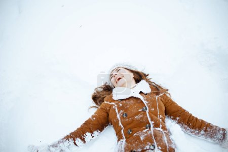 Téléchargez les photos : Femme élégante souriante en chapeau marron et écharpe en manteau en peau de mouton faisant des anges de la neige à l'extérieur de la ville en hiver. - en image libre de droit