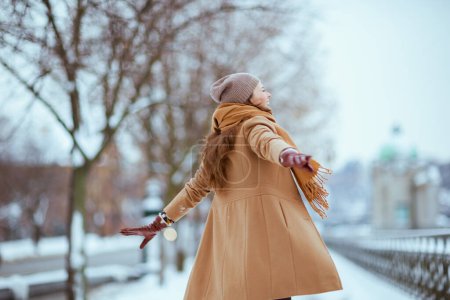 Foto de Visto desde atrás mujer en sombrero marrón y bufanda en abrigo de camello con guantes y brazos levantados regocijándose afuera en la ciudad en invierno. - Imagen libre de derechos