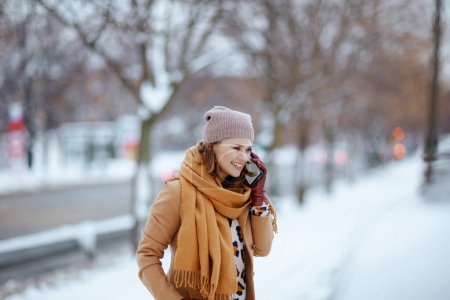 Foto de Mujer elegante sonriente en sombrero marrón y bufanda en abrigo de camello con guantes hablando en un teléfono inteligente fuera de la ciudad en invierno. - Imagen libre de derechos