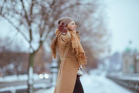 Photo for Smiling modern 40 years old woman in brown hat and scarf in camel coat with gloves rejoicing outside in the city in winter. - Royalty Free Image