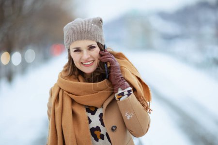 Foto de Sonriente elegante mujer de 40 años con sombrero marrón y bufanda con abrigo de camello con guantes usando un teléfono inteligente afuera en la ciudad en invierno. - Imagen libre de derechos