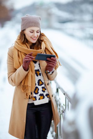 Photo for Happy modern 40 years old woman in brown hat and scarf in camel coat with gloves sending text message using smartphone outside in the city in winter. - Royalty Free Image