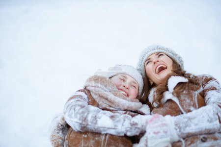 Foto de Sonriente madre y el niño con estilo en abrigo, sombrero, bufanda y manoplas que ponen en la nieve al aire libre en el parque de la ciudad en invierno. - Imagen libre de derechos