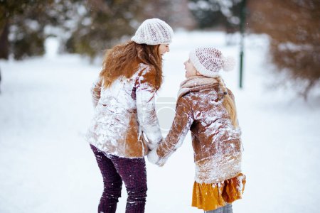 Photo for Happy modern mother and daughter in coat, hat, scarf and mittens in snowy clothes outdoors in the city park in winter. - Royalty Free Image
