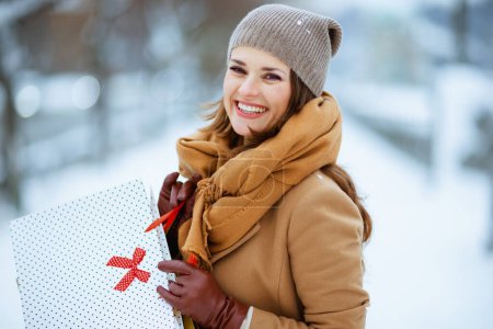 Téléchargez les photos : Heureuse femme élégante en chapeau marron et écharpe en manteau de chameau avec des sacs à provisions à l'extérieur de la ville en hiver. - en image libre de droit