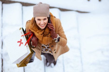 Photo for Upper view of smiling stylish woman in brown hat and scarf in camel coat with gloves and shopping bags speaking on a smartphone outside in the city in winter. - Royalty Free Image