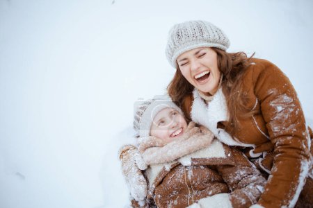 Téléchargez les photos : Mère et fille modernes souriantes en manteau, chapeau, écharpe et mitaines posées dans la neige à l'extérieur dans le parc de la ville en hiver. - en image libre de droit