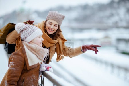 Foto de Sonriendo elegante madre e hija en abrigo, sombrero, bufanda y manoplas con bolsas de compras apuntando a algo al aire libre en la ciudad en invierno. - Imagen libre de derechos