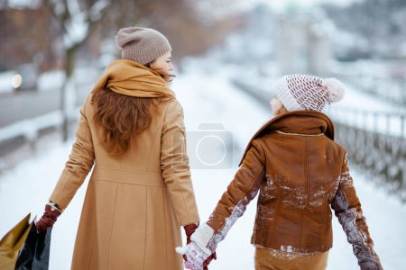 Téléchargez les photos : Vu de derrière élégante mère et fille en manteau, chapeau, écharpe et mitaines marchant à l'extérieur dans la ville en hiver. - en image libre de droit