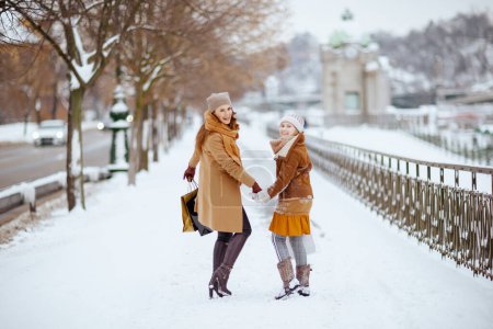 Photo for Full length portrait of smiling modern mother and daughter in coat, hat, scarf and mittens with shopping bags walking outdoors in the city in winter. - Royalty Free Image