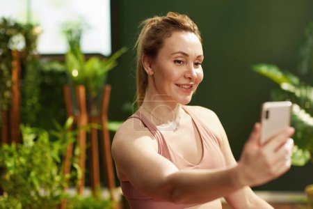 Téléchargez les photos : Heureuse femme au foyer moderne de 30 ans dans le salon vert moderne répondant à l'appel vidéo entrant. - en image libre de droit