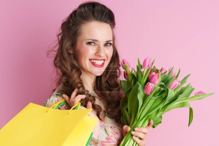 Photo for Smiling stylish middle aged woman with long wavy brunette hair with tulips bouquet and shopping bags isolated on pink. - Royalty Free Image