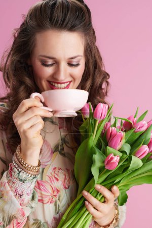 Photo for Smiling stylish 40 years old woman in floral dress with tulips bouquet and cup of tea isolated on pink. - Royalty Free Image