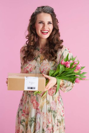 Téléchargez les photos : Femme souriante de 40 ans avec de longs cheveux bruns ondulés avec bouquet de tulipes et colis sur fond rose. - en image libre de droit