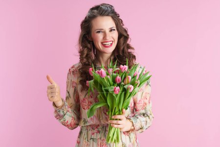 Téléchargez les photos : Heureux élégant femelle avec de longs cheveux bruns ondulés avec bouquet de tulipes montrant pouces isolés sur rose. - en image libre de droit