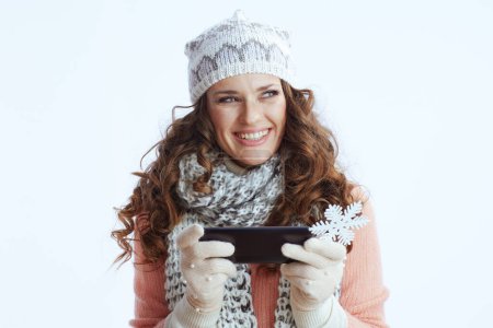 Photo for Hello winter. happy trendy 40 years old woman in sweater, mittens, hat and scarf against white background with snowflake sending text message using smartphone. - Royalty Free Image
