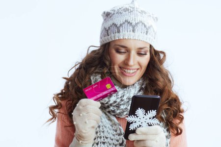 Téléchargez les photos : Bonjour l'hiver. femme moderne souriante en pull, mitaines, chapeau et écharpe sur fond blanc avec flocon de neige, smartphone et carte de crédit faisant des achats en ligne sur le site e-commerce. - en image libre de droit