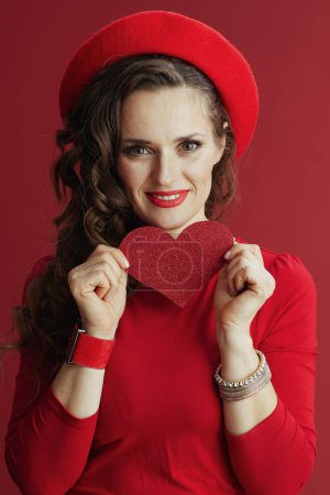 Photo for Happy Valentine. smiling elegant 40 years old woman in red dress and beret against red background with red heart. - Royalty Free Image