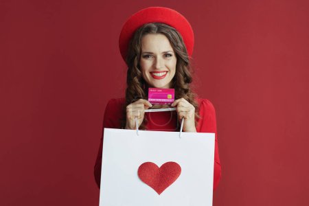 Photo for Happy Valentine. happy elegant female in red dress and beret isolated on red background with credit card and shopping bag and heart. - Royalty Free Image