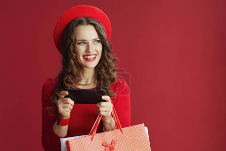 Téléchargez les photos : Joyeux Valentin. heureuse femme moderne avec de longs cheveux ondulés en robe rouge et béret sur fond rouge avec des sacs à provisions en utilisant des applications smartphone et acheter en ligne. - en image libre de droit