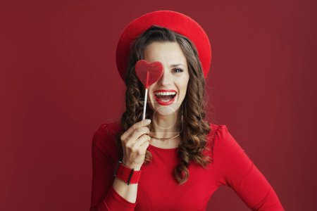 Photo for Happy Valentine. happy stylish middle aged woman with long wavy hair in red dress and beret with heart shaped candy on stick isolated on red. - Royalty Free Image