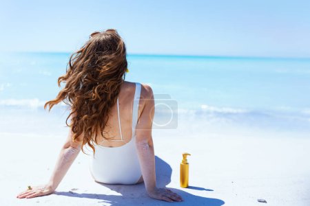 Foto de Visto desde atrás mujer con estilo con protector solar en traje de baño blanco sentado en la playa. - Imagen libre de derechos
