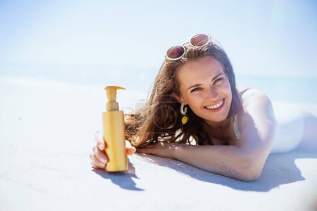 Téléchargez les photos : Portrait de femme heureuse et élégante avec écran solaire en maillot de bain blanc posé à la plage. - en image libre de droit