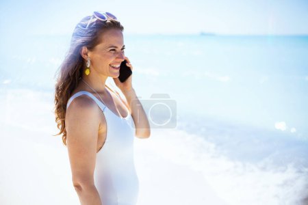 Photo for Happy modern 40 years old woman at the beach in white beachwear speaking on a smartphone. - Royalty Free Image