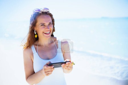 Foto de Feliz mujer con estilo en la playa en traje de baño blanco el envío de mensajes de texto utilizando el teléfono inteligente. - Imagen libre de derechos