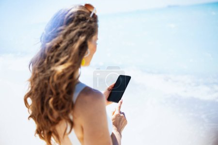 Téléchargez les photos : Vu de derrière femelle moderne à la plage à l'aide d'applications smartphone. - en image libre de droit