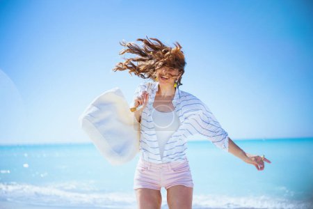 Téléchargez les photos : Femme moderne souriante avec sac en paille blanche en chemise à rayures blanches et short à la plage. - en image libre de droit