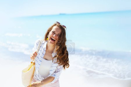 Foto de Sonriente mujer elegante con bolsa de paja blanca que se divierte en la playa. - Imagen libre de derechos