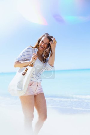 Foto de Mujer moderna feliz con bolsa de paja blanca en camisa blanca a rayas y pantalones cortos en la playa. - Imagen libre de derechos
