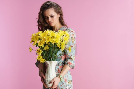 Téléchargez les photos : Femme à la mode en robe florale avec des fleurs de chrysanthèmes jaunes dans un vase isolé sur fond rose. - en image libre de droit