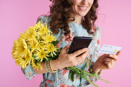 Foto de Primer plano de la mujer moderna feliz en vestido floral con flores de crisantemos amarillos y tarjeta de crédito con aplicación de teléfono inteligente aislado en el fondo rosa. - Imagen libre de derechos