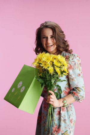Foto de Sonriente elegante mujer de mediana edad con el pelo largo y ondulado morena con flores de crisantemos amarillos y bolsa de compras verde aislado en rosa. - Imagen libre de derechos