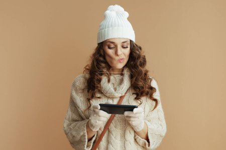 Téléchargez les photos : Bonjour l'hiver. chère femme tendance en pull beige, mitaines et chapeau sur fond beige en utilisant des applications smartphone. - en image libre de droit